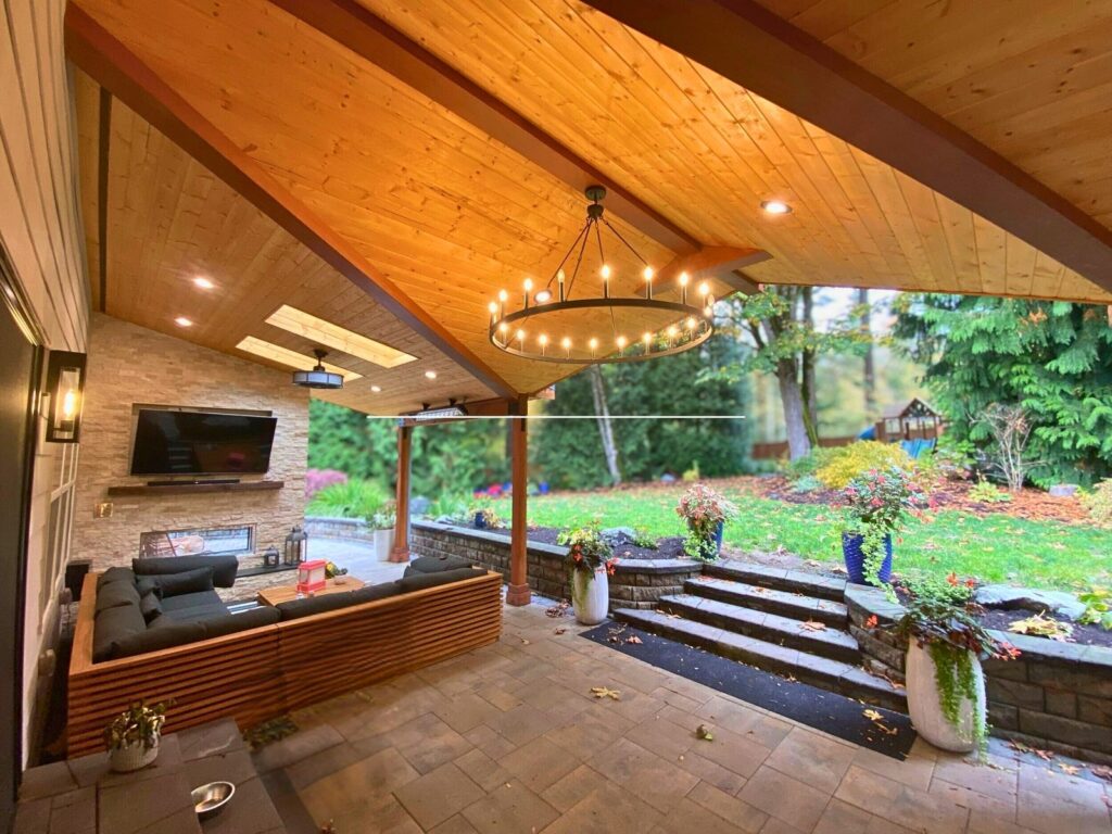 Covered outdoor living room with timber ceiling, chandelier, stone fireplace, and paver patio in the Pacific Northwest