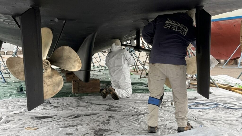Tenhulzen crew under a boat hull on jack stands, with twin bronze propellers and fresh bottom paint