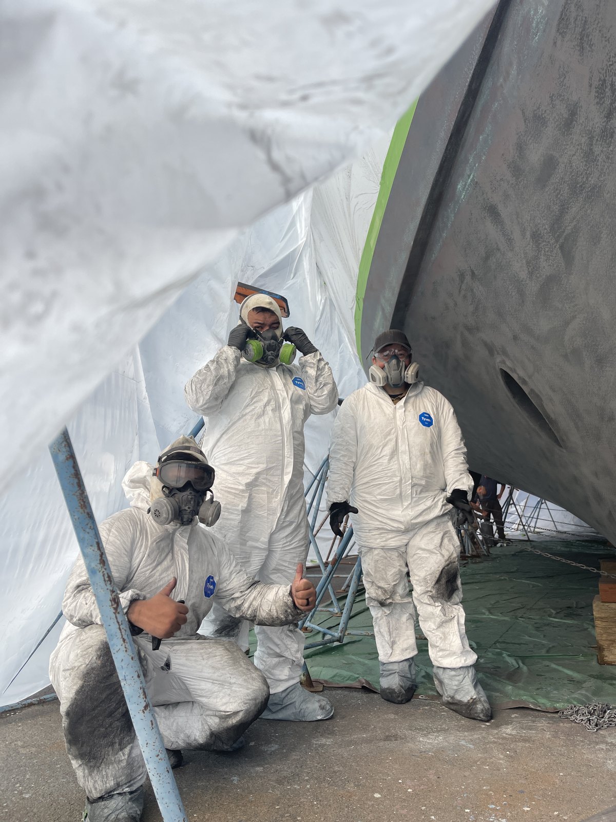 Three Tenhulzen painters in protective suits and respirators under the hull of a boat during bottom paint work